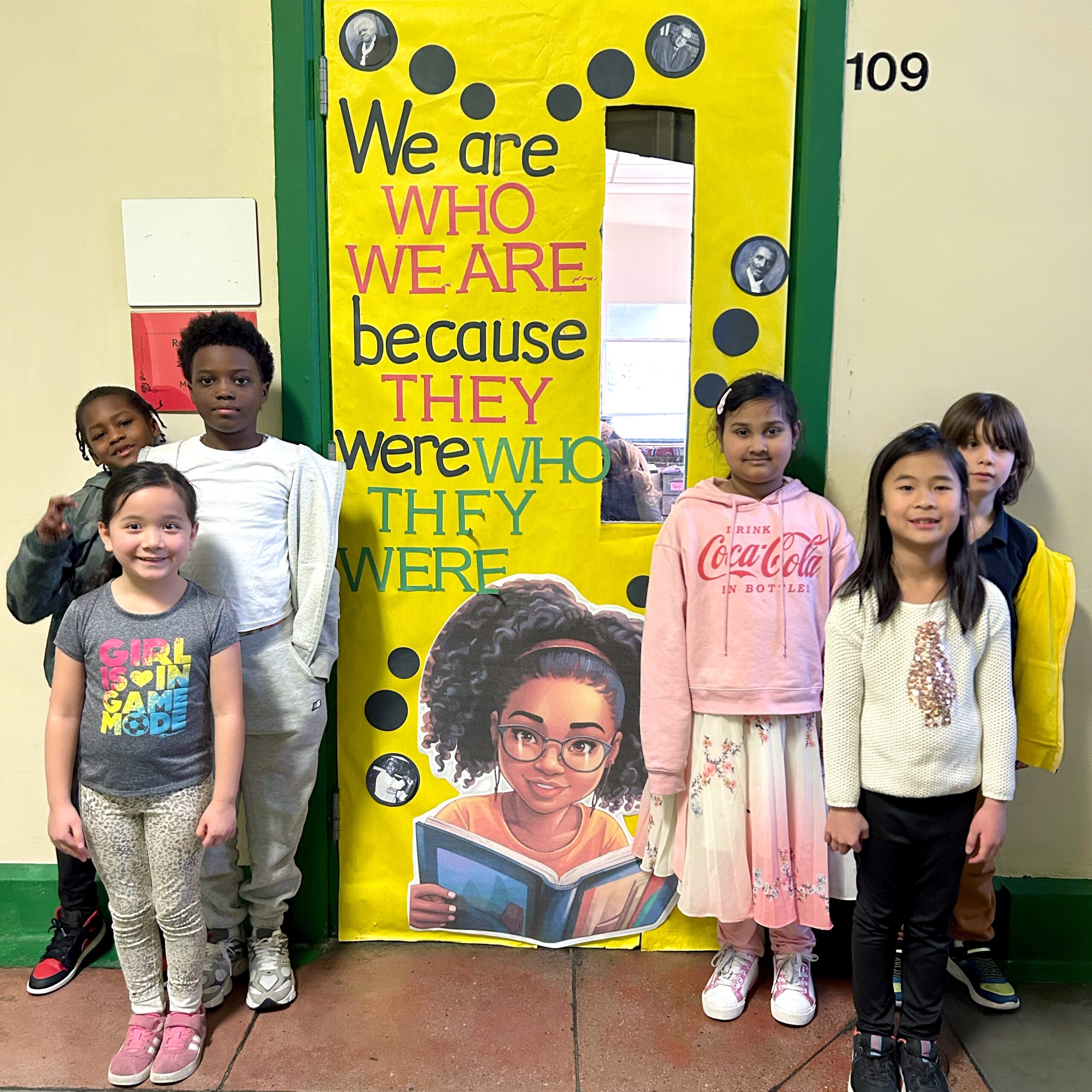 Six young students standing proudly in front of a vibrant yellow classroom door decorated for Black History Month. The display reads, “We are who we are because they were who they were,” and features portraits, polka dots, and an illustrated child reading a book.