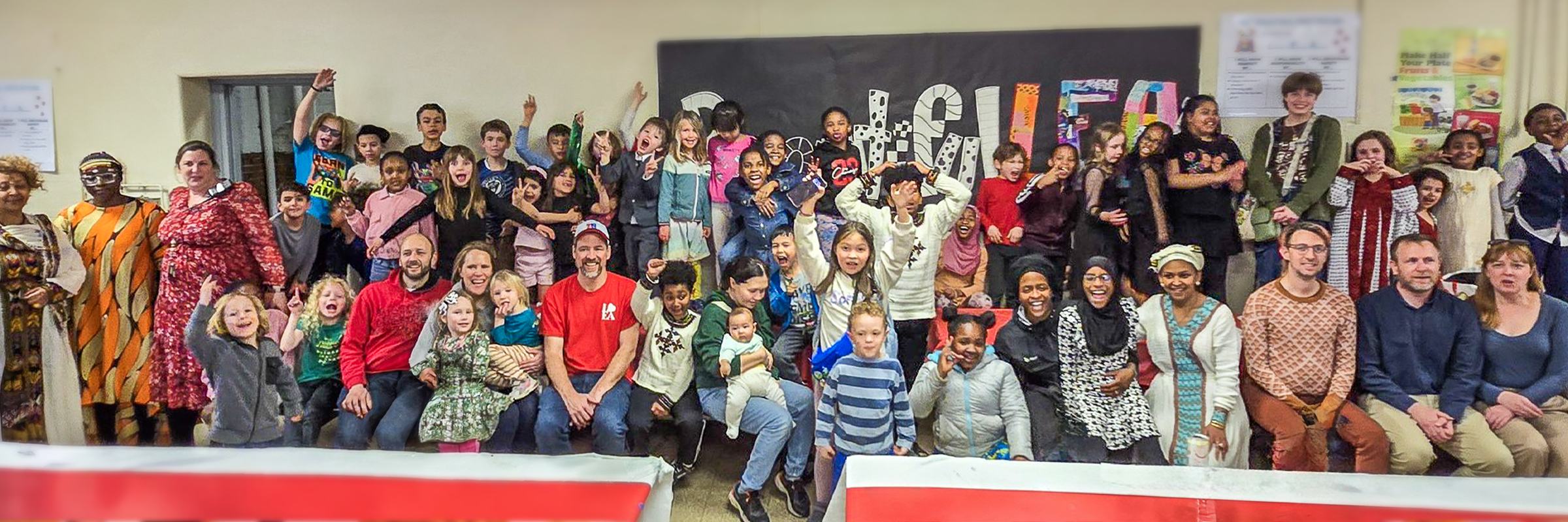 A large, joyful group of children, families, and staff from Henry C. Lea School gathered together in a colorful, multicultural celebration. Participants of all ages are smiling, waving, and posing in front of a decorated wall that reads “Beautiful Faces.”