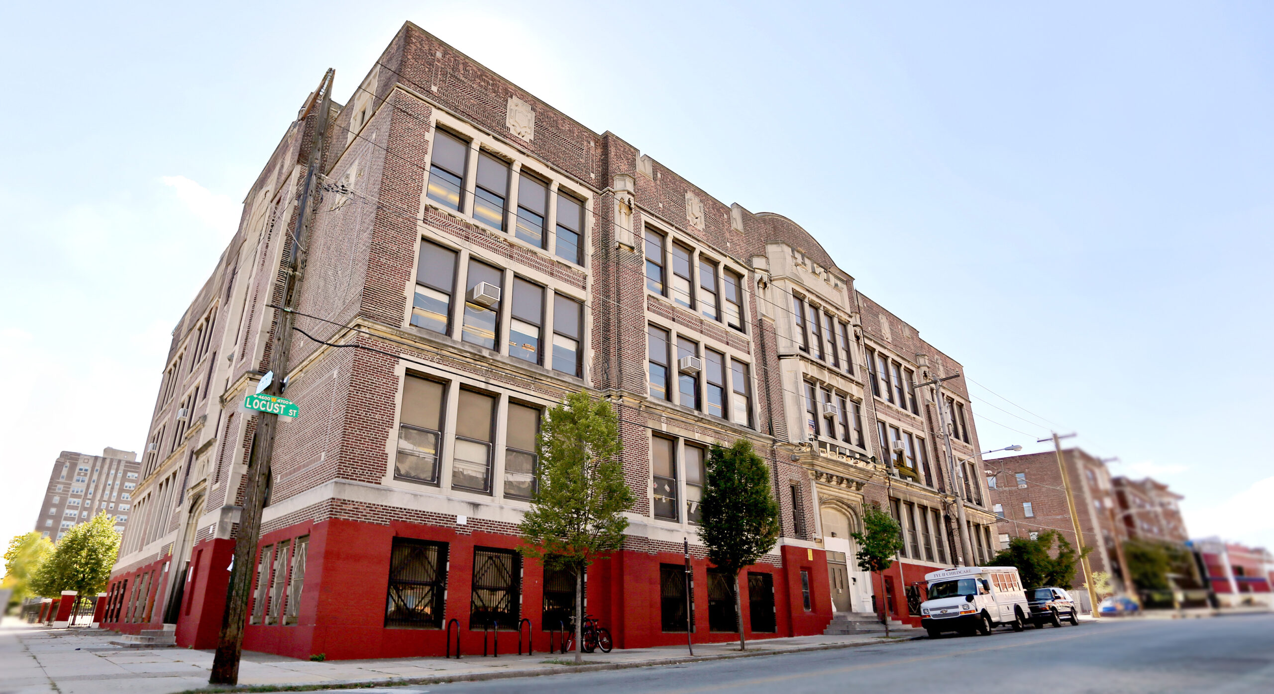 Exterior of the historic Henry C. Lea School building, a large brick structure with red trim, located at the corner of Locust Street in West Philadelphia, with trees and parked cars lining the sidewalk.