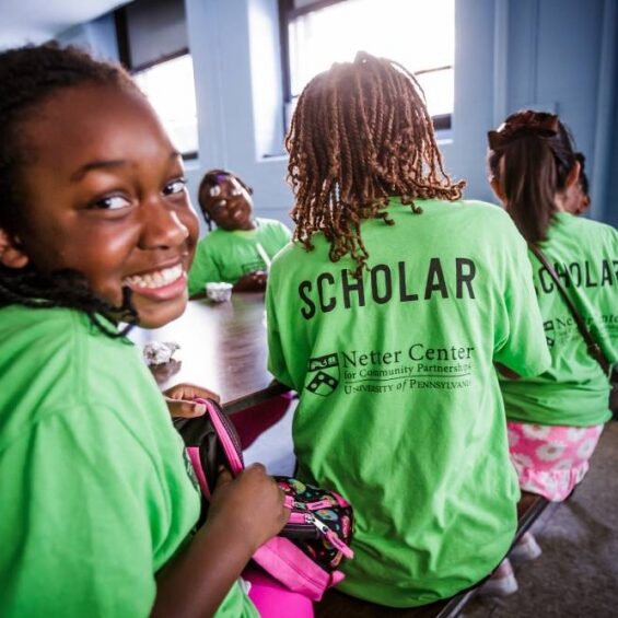 Smiling student in a bright green "Scholar" T-shirt seated at a cafeteria table with peers during the Lea-Penn Summer Scholars program, supported by the Netter Center for Community Partnerships at the University of Pennsylvania.