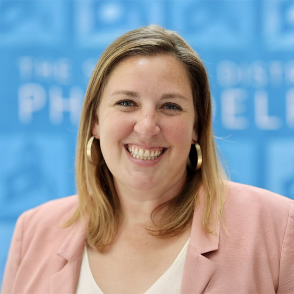 Julia Snyder, Principal of Henry C. Lea School, smiling in front of a blue School District of Philadelphia backdrop, wearing a pink blazer and gold hoop earrings.