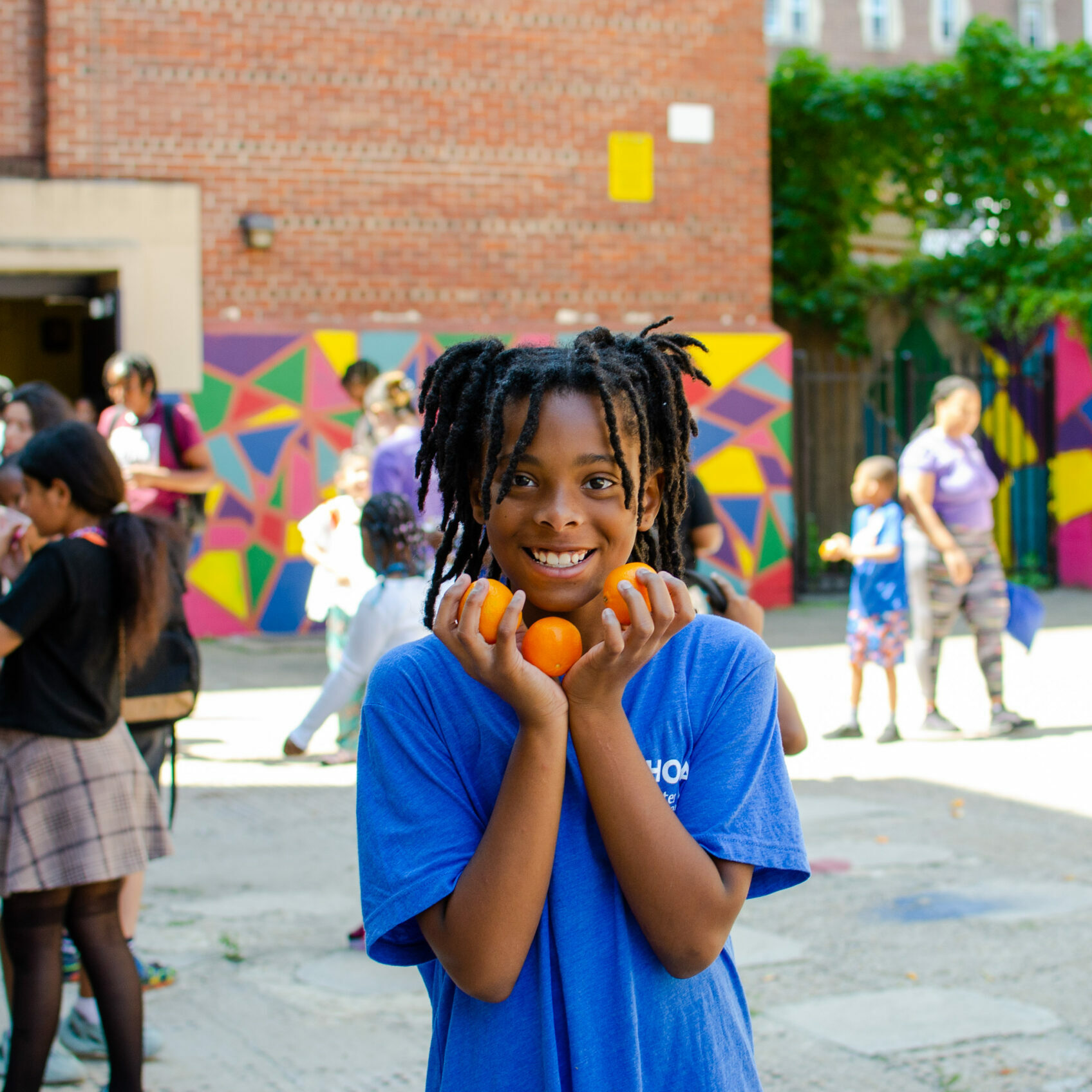Smiling student holding bright orange tomatoes in both hands during an outdoor activity at Henry C. Lea School's UACS summer program. Children and families are gathered in the background near a colorful mural wall.